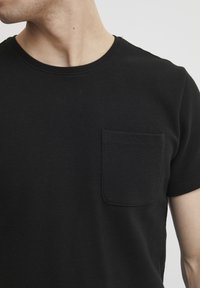 Sort ribbet kortærmet t-shirt med rund halsudskæring og en lomme på venstre bryst. Materialet virker blødt og tekstureret.
