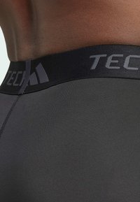 Schwarze Sportshorts mit glatter Textur und einem elastischen Bund, der das graue "TECH"-Logo trägt. Nahaufnahme, die sich auf den Taillenbereich konzentriert.