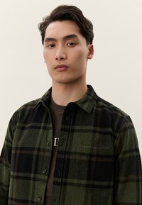 Jaunas vyras su trumpais tamsiais plaukais, dėvintis žalią ir juodą languotą marškinių tipo palaidinę virš tamsiai rudos spalvos marškinėlių, stovintis priešais paprastą šviesią fono sieną.