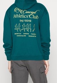 Turkusowa bluza z kapturem z żółtym napisem "Concept Athletics Club" oraz figurami biegaczy, wymieniająca dzielnice Nowego Jorku; osoba w szarych spodniach typu cargo.