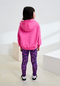 Rosa Kapuzenpullover mit Kordelzug und gerippten Bündchen, kombiniert mit eng anliegenden lila und pink gemusterten Leggings. Schwarze und weiße Turnschuhe.