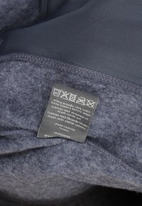 Pflegeetikett auf dunklem Fleecegewebe zeigt Wasch- und Trocknungssymbole, Anweisungen zum Reißverschluss und Waschen mit ähnlichen Farben.