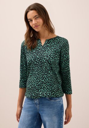 Femme portant un haut à motifs noir et vert à manches longues avec des manches trois quarts et un jean bleu, se tenant devant un arrière-plan neutre.