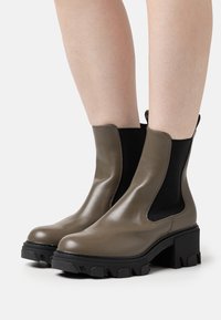 Un par de botines topo con paneles laterales negros de elástico y suelas gruesas negras, llevados sobre piernas desnudas.