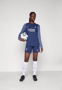 Nike fotbaldrakt i marineblått og lyseblått. Har lange ermer, shorts og laglogo. Spilleren holder en mønstrete fotball. Sorte fotballsko og hvite sokker.