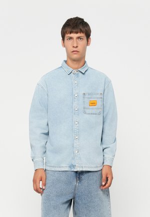 Light blue denim shirt with a button-up front, pointed collar, and a chest pocket featuring a yellow patch label. Textured fabric.