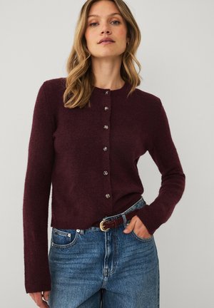 Kvinde iført en mørk burgunderfarvet cardigan med knapper og lange ærmer samt blå jeans med brun bælte, står med den ene hånd i lommen.