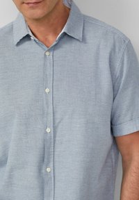 Camicia a maniche corte con bottoni di colore azzurro chiaro, con un motivo testurizzato, dotata di colletto e bottoni bianchi sulla parte anteriore.