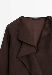 Ruskea, teksturoitu takki, jossa on valuva kaulus ja syvä V-nuora muotoilu. Kankaan pinnassa on hienoinen kiilto, joka lisää ulkonäön eleganttia ilmettä.