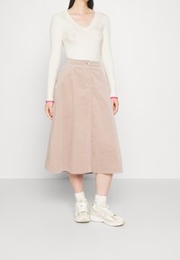 Vrouw in een witte geribbelde V-hals truien, beige midi corduroy rok, witte sokken en beige New Balance sneakers, staande tegen een witte muur.