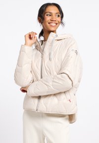 Beige gewatteerde pufferjack met een capuchon. Beschikt over een rits aan de voorkant, grijze trekkoorden en subtiele stiksels op de stof.
