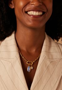 Collier en chaîne dorée avec une pierre bleue ovale et un pendentif en cristal transparent. Porté sur un blazer à rayures fines de couleur claire.