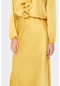 Persona che indossa un vestito lungo giallo in satin con maniche a sbuffo e un dettaglio a ruffle vicino al collo, con le mani appoggiate ai lati.