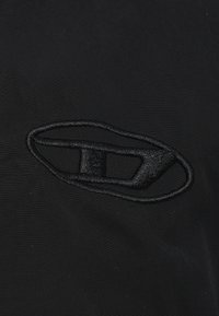 Melns audums ar teksturētu virsmu, centrā atrodas izteiksmīgs melns izšūts ovāls logotips, kas atspoguļo minimālistisku dizainu.