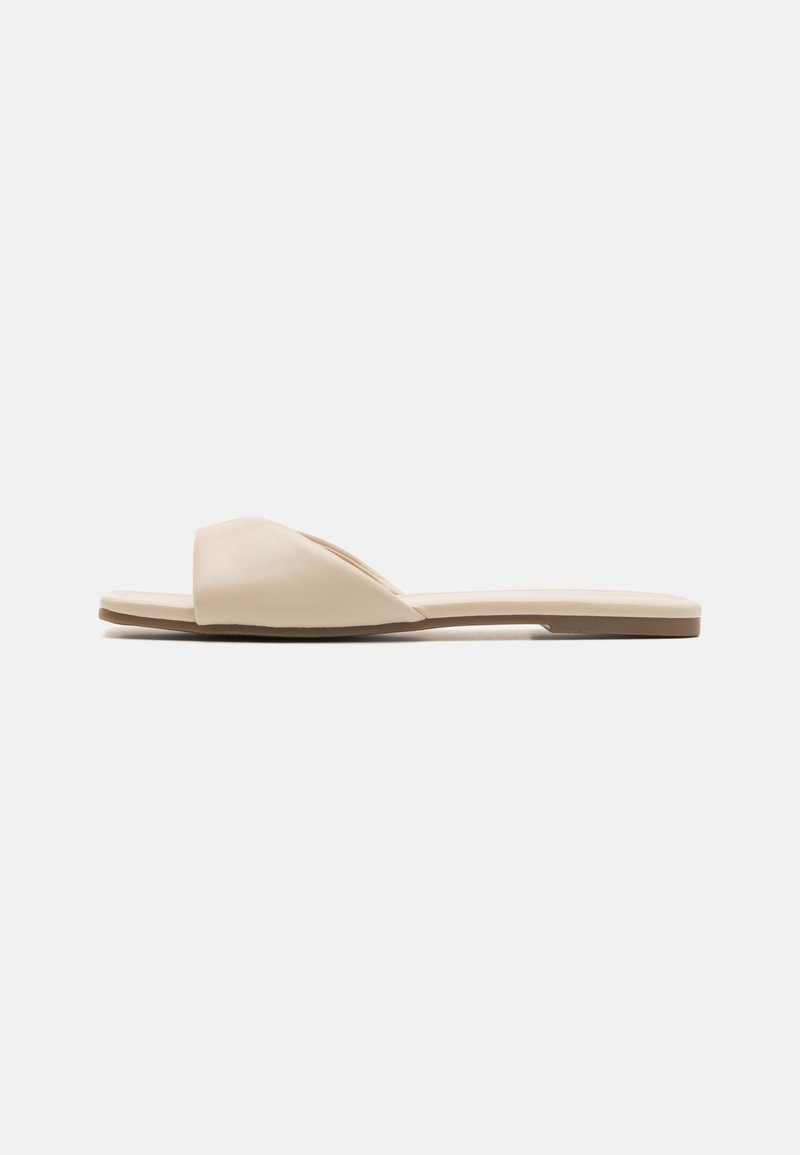 Sandales slides beiges avec une bride souple et incurvée, semelle plate, présentant une texture lisse et un style minimaliste.