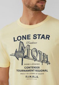 Geel katoenen T-shirt met een grafische print met "LONE STAR MOTEL" en verschillende tekstelementen in donkerblauw, ronde halslijn, korte mouwen.