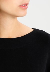 Top nero a maglia rigata con scollatura ampia e tonda. Il tessuto sembra morbido con una leggera texture. Inqudratura ravvicinata del colletto e della zona delle spalle.