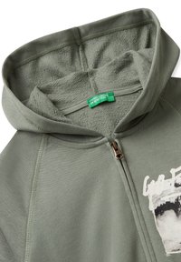 Sweatshirt à capuche vert olive, avec une fermeture éclair, texture douce et un motif imprimé sur le devant. Étiquette visible à l'intérieur du col.