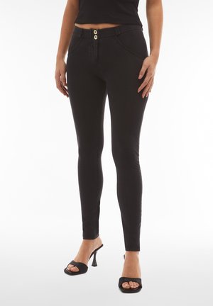 Nainen pukeutuneena mustiin korkeavyötäröisiin kapealahkeisiin farkkuihin, joissa on kaksinappinen suljenta, ja mustiin avokkaisiin korollisiin sandaaleihin, seisoo valkoista taustaa vasten.