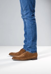 Brune ruskindssko med glat tekstur og lukket snørebånd, kombineret med blå denimjeans, vist fra siden mod en grå baggrund.