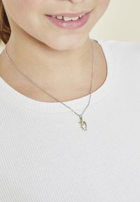 Silverhalsband med en liten enhörningsberlock med en gul accent. Bäraren har på sig en ribbad vit skjorta som tydligt visar halsbandet.