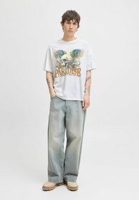 Hvid grafisk t-shirt med et flerfarvet kranium og vinger design, parret med løstsiddende lyse jeans og beige fodtøj.