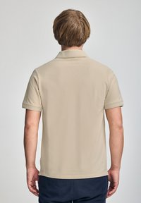 Camisa polo de manga corta beige con una textura suave, cuello plegado y dobladillo recto, vista desde la parte posterior. Diseño sencillo, sin patrones.