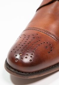 Chaussure en cuir brun avec un bout arrondi, présentant des perforations et des coutures discrètes. Texture lisse avec un fini légèrement brillant.