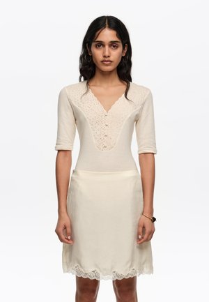 Mujer con un top de punto color crema con escote en V con borde festoneado y botones, combinado con una falda de satén crema con ribete de encaje en el dobladillo.