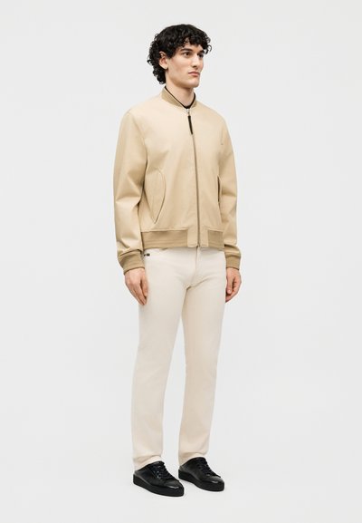 Fekete göndör hajú férfi bézs bomber dzsekiben, törtfehér nadrágban és fekete sportcipőben, egyszerű világosszürke háttér előtt áll.