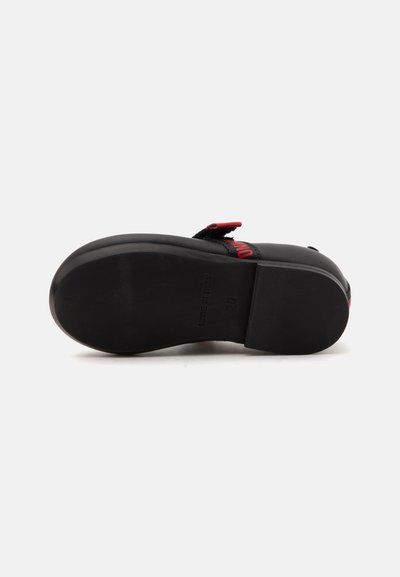 Semelle de chaussure face vers le haut, noire avec des bords lisses et arrondis, montrant une bande velcro et un détail rouge sur la partie supérieure.