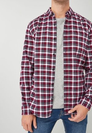 Man draagt een rood-wit geruit overhemd over een grijs T-shirt en blauwe spijkerbroek, hand past de zoom van het overhemd aan.