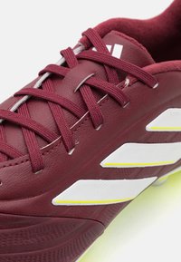 Botín de fútbol color burdeos con parte superior de cuero texturizado, diseño de tres rayas en blanco y detalles amarillos en la suela. Cuenta con cordones trenzados.