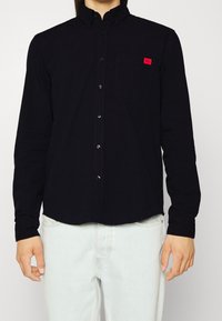 Camisa de botones de manga larga en negro, mezcla de algodón, con un bolsillo en el pecho y un pequeño parche de logo rojo. Textura suave y cuello clásico.