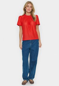 Top rosso a maniche corte con scollatura arricciata e motivo testurizzato, abbinato a jeans blu a gamba dritta e ballerine con stampa leopardata.