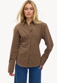 Chemise à carreaux marron à manches longues avec col, poche avant et patte de boutonnage, portée avec un jean en denim bleu taille haute.