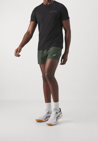 Camiseta negra de manga corta de ASICS, pantalones cortos verdes oliva, zapatillas deportivas blancas y negras con acentos naranjas, de pie sobre un fondo neutro.
