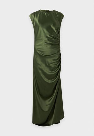 Une femme porte une robe longue jusqu’au sol en satin vert olive, froncée sur les côtés, et des escarpins noirs à brides, sur un fond blanc uni.