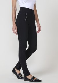 Visoko pasne črne legice iz raztegljive tkanine, z okrasnimi gumbi na sprednjih žepih in slim-fit dizajnom.