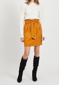 Jupe plissée orange avec un détail de taille nouée, associée à un haut crème à manches longues et des bottes noires molles montant jusqu'au genou.