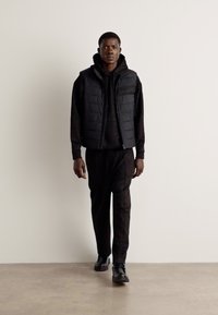 Black padded vest over a black hoodie, paired with dark sweatpants and black shoes. Smooth texture, sleek design, and minimalistic details.