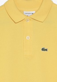 Gul polo skjorta i texturerat tyg med ribbad krage, treknappsstängning och en grön krokodilslogotyp på bröstet.