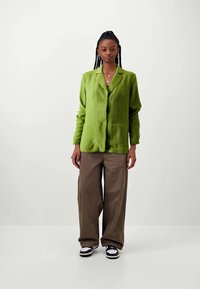 Green textured button-up jacket with a collar, paired with loose brown pants and black and white sneakers. Neutral background.