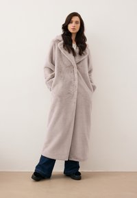 Długi, jasnoszary sztuczny płaszcz z szalowym kołnierzem, z bocznymi kieszeniami i luźnym krojem, noszony na ciemnoniebieskie szerokie spodnie.