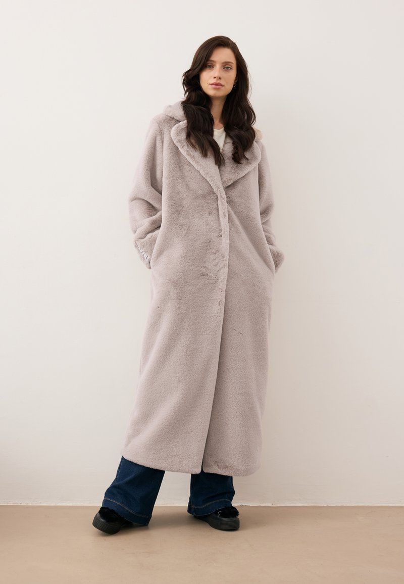 Długi, jasnoszary sztuczny płaszcz z szalowym kołnierzem, z bocznymi kieszeniami i luźnym krojem, noszony na ciemnoniebieskie szerokie spodnie.