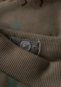 Bruin brei stof met ribbelstructuur, met een iets lichtere ribbelkraag. Een doorzichtige knop met een bloemdessin is dichtbij de kraag genaaid.