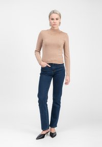 Mujer de pie contra un fondo blanco, vistiendo un suéter beige de manga larga con adornos, jeans azul oscuro y tacones negros con punta.