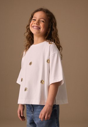 Junges Mädchen mit lockigem Haar lächelnd, trägt ein weißes, lockeres Hemd mit goldenen Pailletten-Blumenmustern und blaue Jeans vor einfachem Hintergrund.