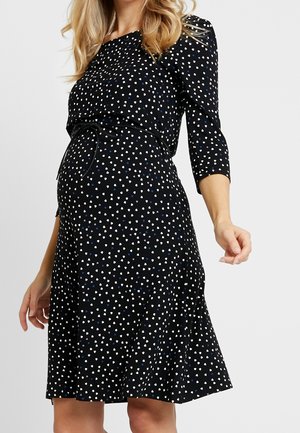 Kobieta w ciąży ubrana w czarną sukienkę do kolan w białe i niebieskie wzory serc, z rękawami do łokci, pozująca na jednolitym tle.