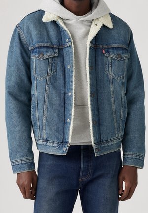 Mand iført en blå denimjakke med hvid fleeceforing over en lysegrå hættetrøje og mørkeblå jeans, stående mod en ensfarvet baggrund.